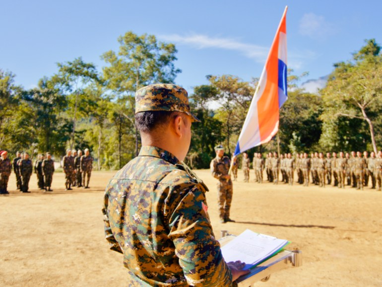 A Chin officer holding a clip board at a roll call with a red, white and blue flag at the centre of the parade ground