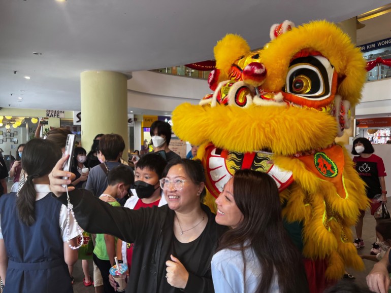 Two women take a selfie with the lion after the performance. They are smiling and look very happy. The lion is yellow with furry eyes and upper lip and painted with gold, red and black