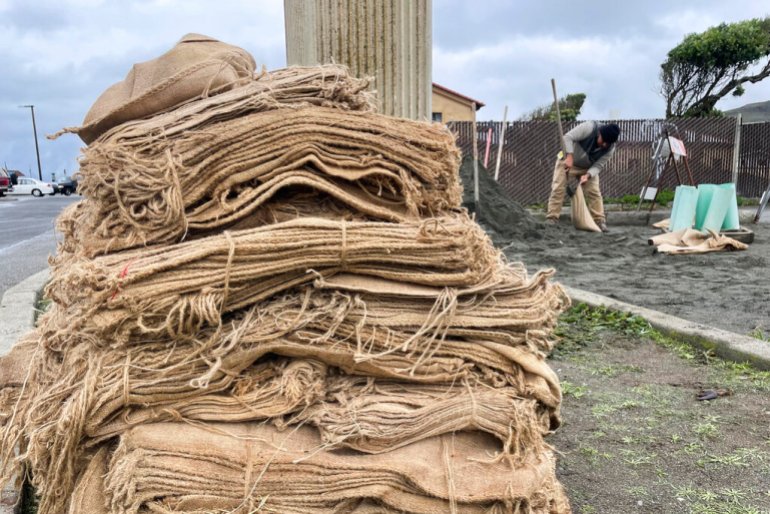 A stack of empty sandbags. Behind them, to the right, a map is digging is holding one sandbag and filling it with dark sand