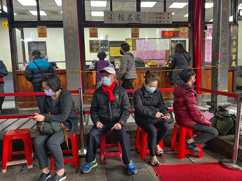 People waiting to an tan sui or pray to other gods at the temple.  They are sitting on red plastic stools.  Behind them are counters where they pay for lamps and candles