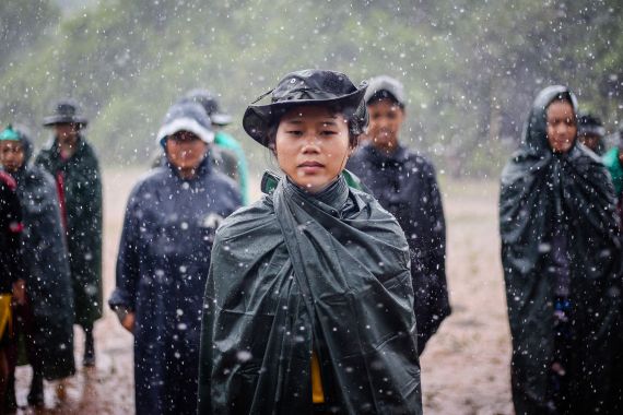 A Karenni female fighter standing with her comrades in the rain. She is wearing a camouflage wide-brimmed hat and a plastic mac. She looks determined