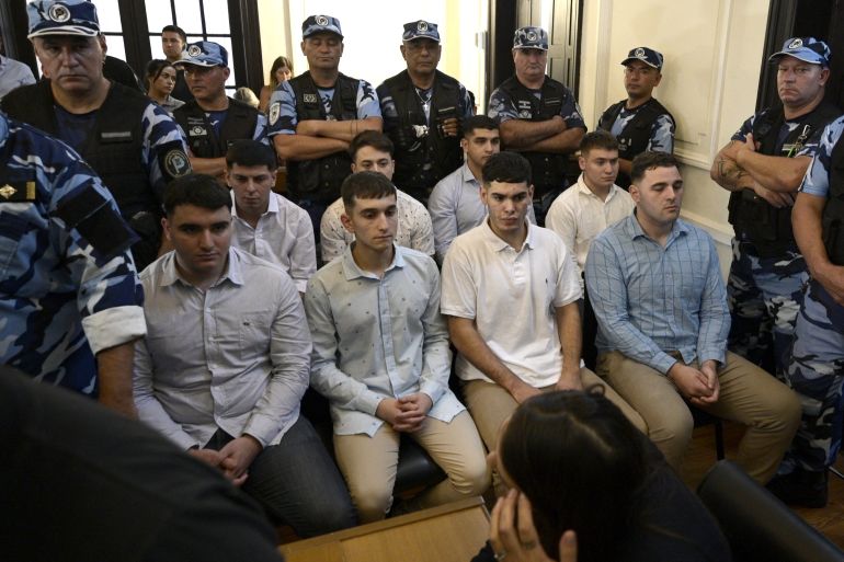 Two rows of young men in white or light blue collared shirts sit with hands folded in a courtroom, as they hear the verdict surrounded by police and onlookers