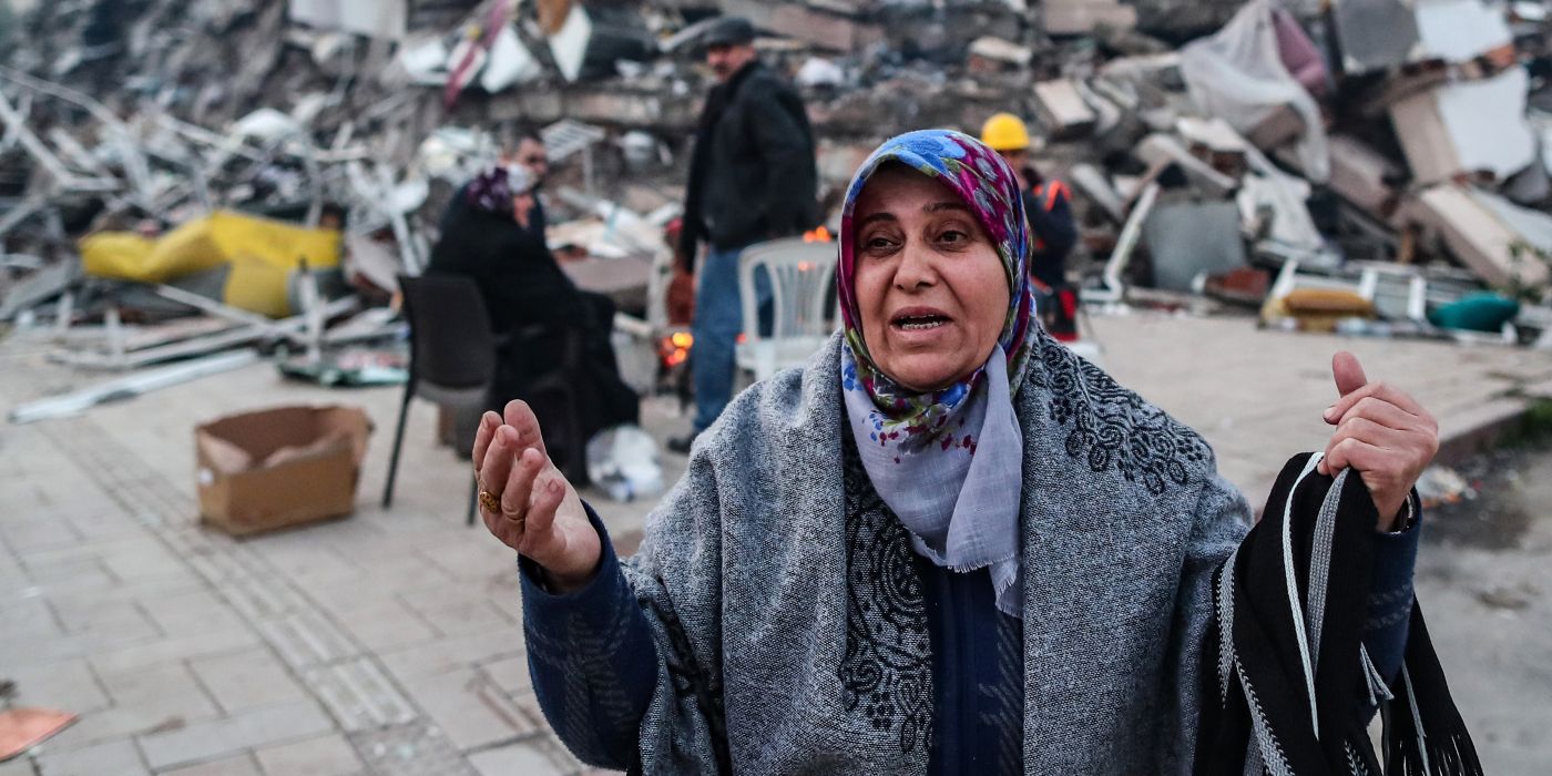 A woman reacts as others warm up around a fire in front of a collapsed building following a major earthquake in Hatay, Turkey