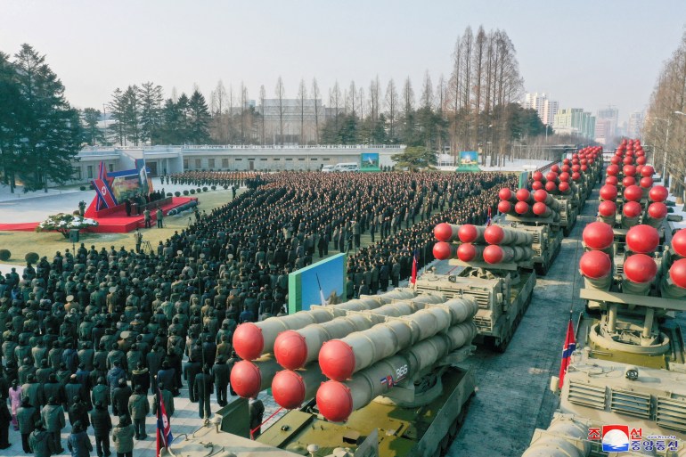 A large group of men are watching Kim Jong Un speak at a special stage with a double row of super-large rocket launchers lined up on the road behind them. There are trees in the background and high-rise buildings in the distance.