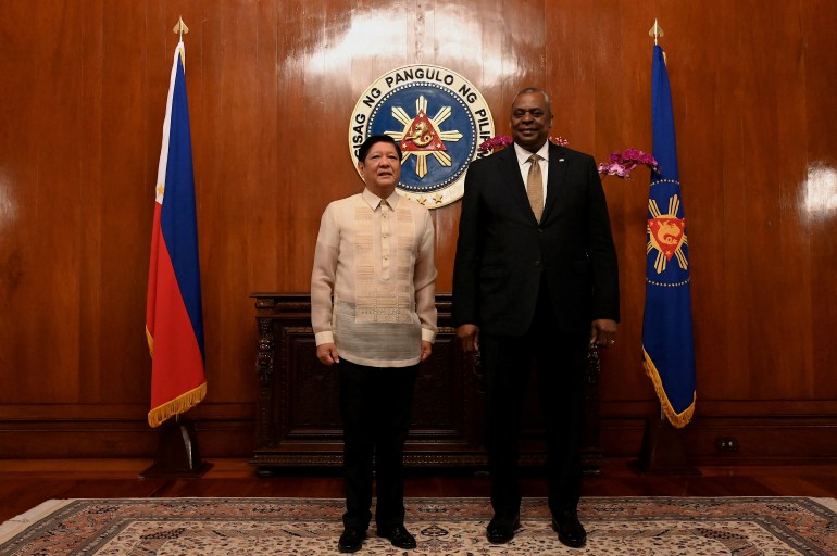 Ferdinand Marcos Jr and Lloyd Austin standing next to each other at the Malacanang Palace.  Marcos Jr is wearing a barong, a traditional Filipino shirt, and Austin is wearing a dark suit.  Behind them are the presidential seal and the flags of the two countries