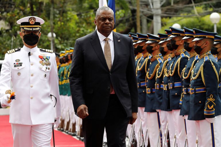 US Defense Secretary Lloyd Austin walks past an honour guard at Camp Aguinaldo in Quezon City. His is accompanied by an officer in a white uniform. The guard are lined up and their uniforms are green jackets and white trousers. They are wearing face masks.