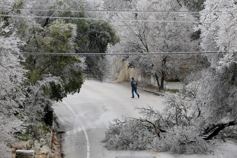 A person walks down an icy road, framed by trees white with frost, one of which has tumbled down onto the pavement