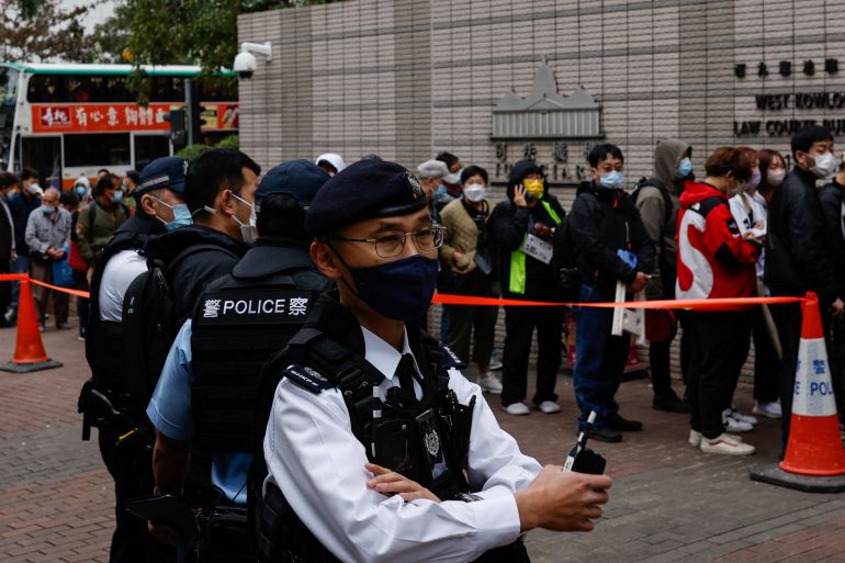 Police outside the West Kowloon Magistrates' Courts. One is facing the camera. He is wearing a white shirt with black vest and hat, as well as a face mask and is holding a walkie-talkie. Other officers are behind him and there is a queue of people standing behind a red tape to get into the court.