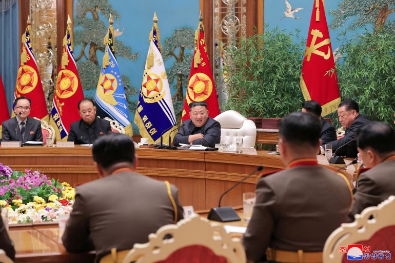 North Korean leader Kim Jong Un at a meeting of the party's military commission. He is sitting at at a circular desk with a large flower arrangement on a table in the middle. There are flags behind him and a painting of trees and birds on the wall. He appears to be laughing. He is sitting on a white upholstered chair and is leaning on his crossed elbows on the desk.