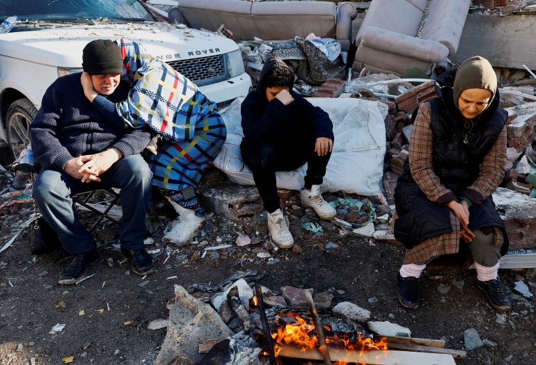 People sit next in front a fire with rubble around them. On person is hugging a man while a boy sits with his head in his hands. A woman looks at the ground in distress.