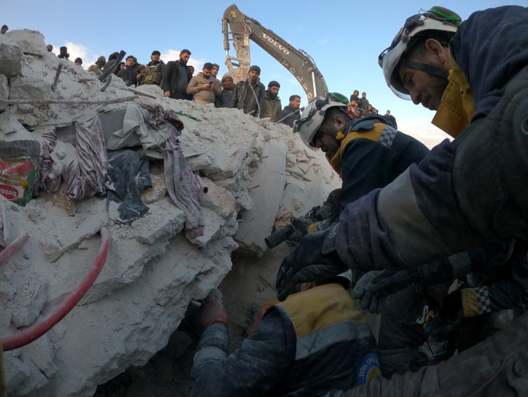 Rescue workers searching a pile of rubble for survivors. There are a group of people standing looking down on what's happening. The outline of an excavator can also be seen at the top 