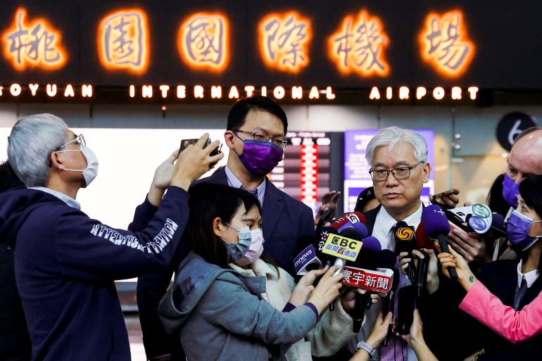 Andrew Hsia surrounded by reporters at the Taipei airport before leaving for China. He has white hair and glasses and is wearing a suit. The journalists are holding microphones in front of him. The airport sign is behind in both English and Chinese characters.