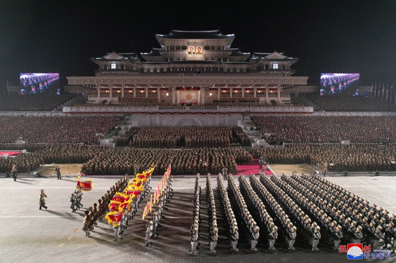 A view from the top shows perfect rows of troops marching behind a leader. Behind the leader and ahead of the troops are what look to be three men, one holding a flag. The stands behind them are filled with people all wearing the same dark green military gear.