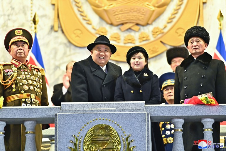 Kim stood smiling on the podium next to her daughter.  Both wore black coats and black hats.  Two men walked beside Kim and her daughter, one wearing a military uniform with a medal.