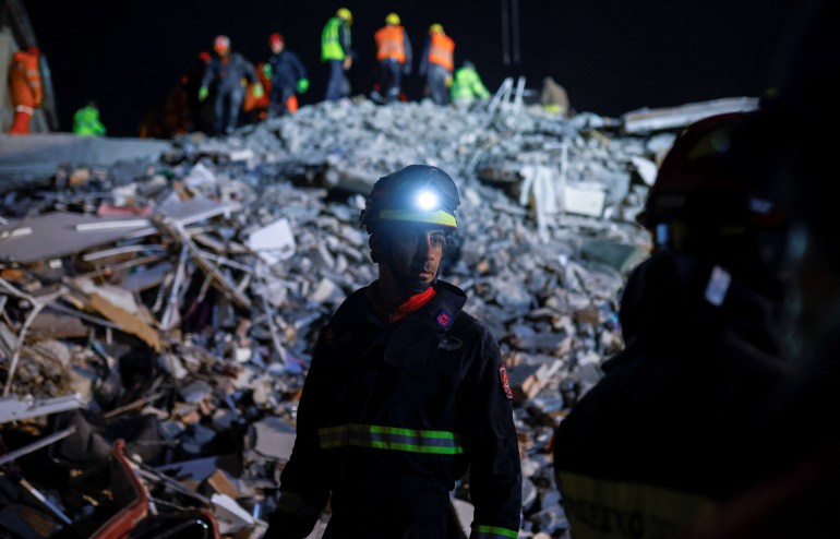 Rescuer wearing a hat with a light stands in front of a hill of rubble with many more rescuers on top of it, in the background. It is nighttime. 