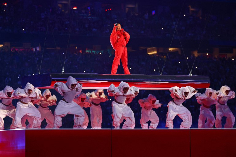 Rihanna descended to the stage on a platform.  He wears a red jumpsuit.  The dancers are on the stage and bent over.  They wear white ski suits.