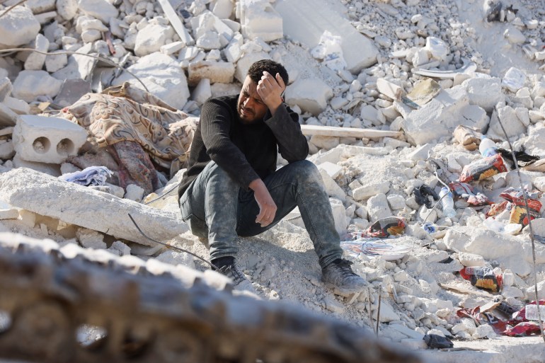 Man is sitting on rubble with his head in his hand, looking devastated