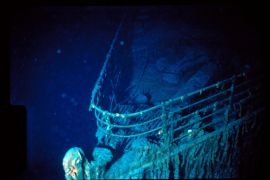 Bow of the Titanic wreck which looks an aqua colour with barnacles on it. The ocean water is a deep, dark blue