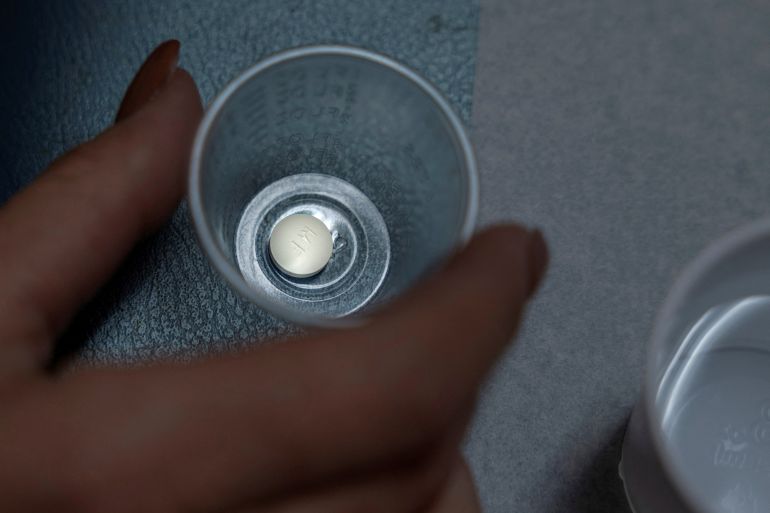 A manicured hand grabs a tiny plastic cup containing a round, white pill