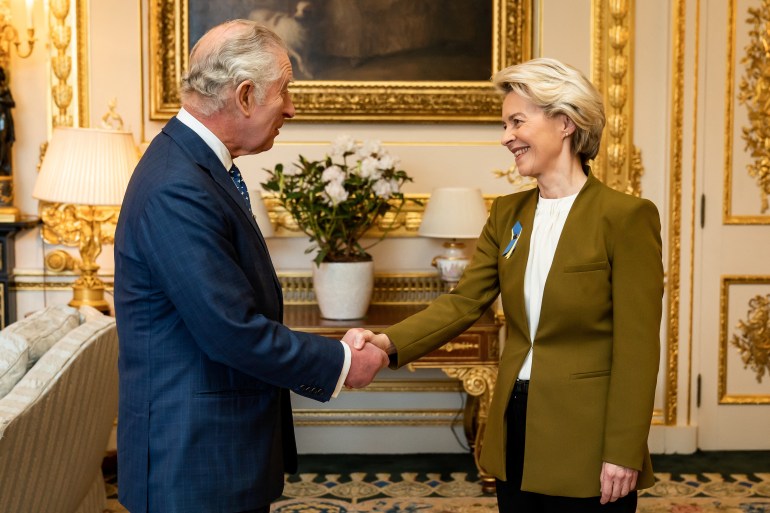 European Commission President Ursula von der Leyen meets King Charles III at Windsor Castle. The two are shaking hands and smiling.  Von der Leyen has a ribbon in the blue and yellow of Ukraine on her lapel. The room has gilded walls and there is a plant with white flowers on the ornate table behind them.  