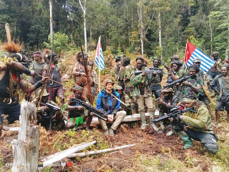 Philip Mehrtens sitting on a log in the forest surrounded by TPN-PB fighters. They have weapons and there are two Papuan independence flags. The fighters have weapons including guns and bows and arrows. Most are in military-type clothing but some are in traditional dress. 