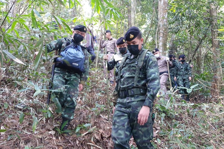 Task force members, some in camouflage uniforms and others in grey, wearing black face masks trudge through a thick forest. Some hold long sticks to help them with the walk. They're wearing army caps.