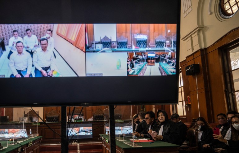 A video screen showing the start of the trial into the Malang football disaster, with the judges on one side and defendants on the other. The accused initially appeared on video link