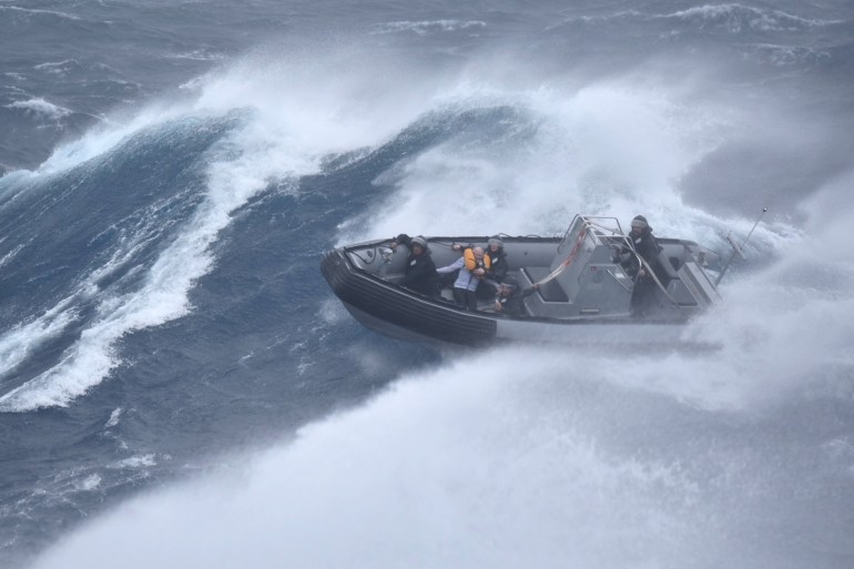 An inflatable dinghy powering across massive waves and spray.  A rescued sailor is on board wearing an orange life vest.  There are also three members of the New Zealand Defense Force on board