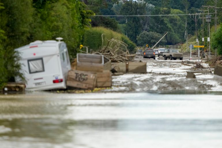 A caravan, tree debris and wooden crates in piles of mud on a road near Napier. There is flooding in the front of the photograph and mud covered trucks in the background