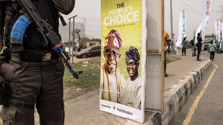A private security guard with an automatic weapon stands near a a road and next to an election poster for All Progressive Congress leader Bola Tinubu in Lagos, Nigeria.