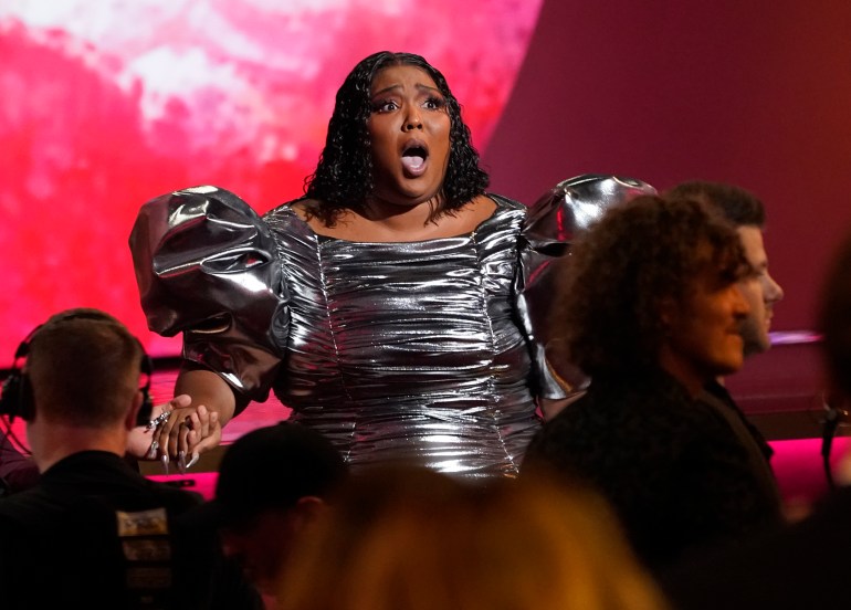 Lizzo receives a Grammy Award for Record of the Year.  She is wearing a dark silver dress with large puffed sleeves, her dark hair is loose.  She looks shocked and squeezes someone's hand.