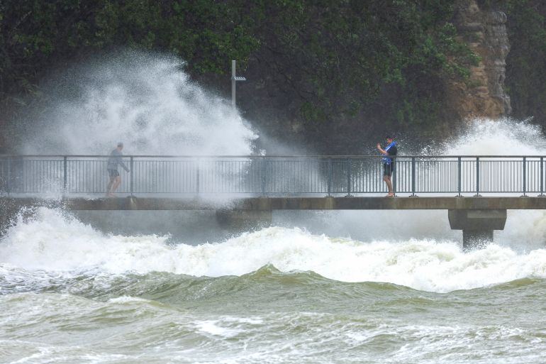 Huge waves crash into cliffs in Auckland. There is a walkway in front where a couple of people can be seen
