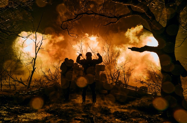 A Ukrainian soldier silhouetted against orange flames after firing artillery at Russian positions near Bakhmut, Ukraine. He has his arms in the air. There are trees at the sides in the foreground and debris behind.