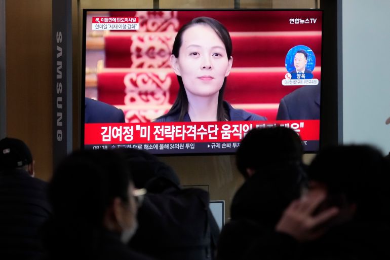 Kim Yo Jong seen on a television screen. She appears to be standing at the bottom of some stairs carpeted in red with a gold pattern. She looks serious.