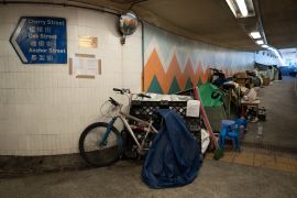 A bicycle propped up against the wall of an underpass next to other belongings of the homeless people who have taken shelter there. There's a blue street sign on the wall and a sing saying no smoking.