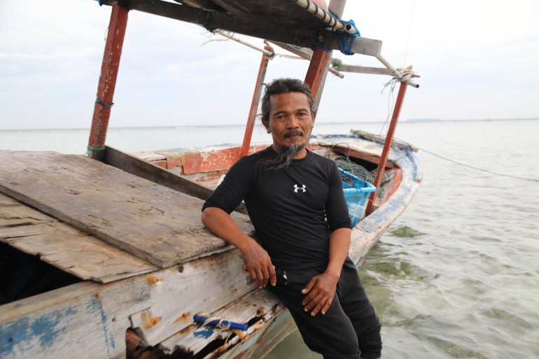 Bobi appuyé contre un bateau de pêche qui est en eau peu profonde.  Il porte un pantalon noir et un t-shirt noir