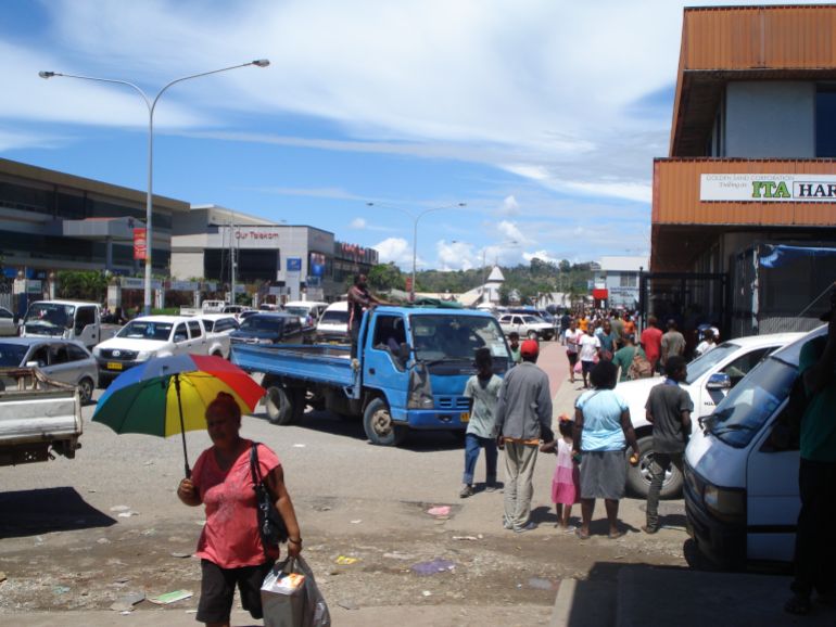 A street in Honiara. There is a woman walking towards the camera holding a large umbrella, crowds walking the other way, and lots of cars and vans. There are also potholes and rubbish.
