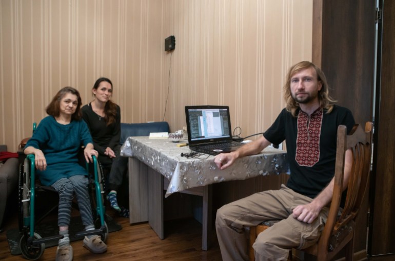 A photo of Oksana’s mother Tanya sitting in a wheelchair with Oksana sitting in a chair next to her and her husband Sergio sitting on a table with a laptop in front of them.