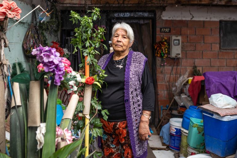 Mireia Godoy, a woman in a black shirt and a long purple vest, stands in a yard in front of her house, surrounded by plants, buckets and coolers
