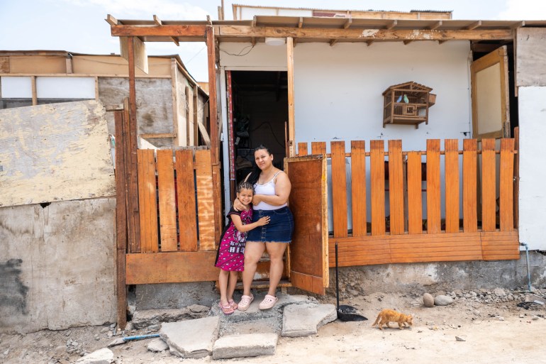 Marian and her daughter pose in front of a makeshift home, which is white with a flat roof and a wooden fence-like structure in the front