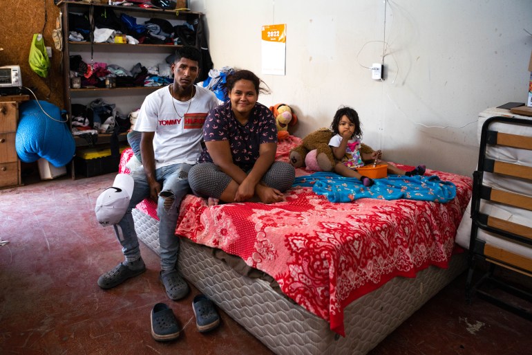 A family of three - a man, a woman and a child - sits on a mattress covered by a red blanket. The little girl is leaning against a brown teddy that is bigger than her. She has an orange bowl of food lying in front of her on the bed.