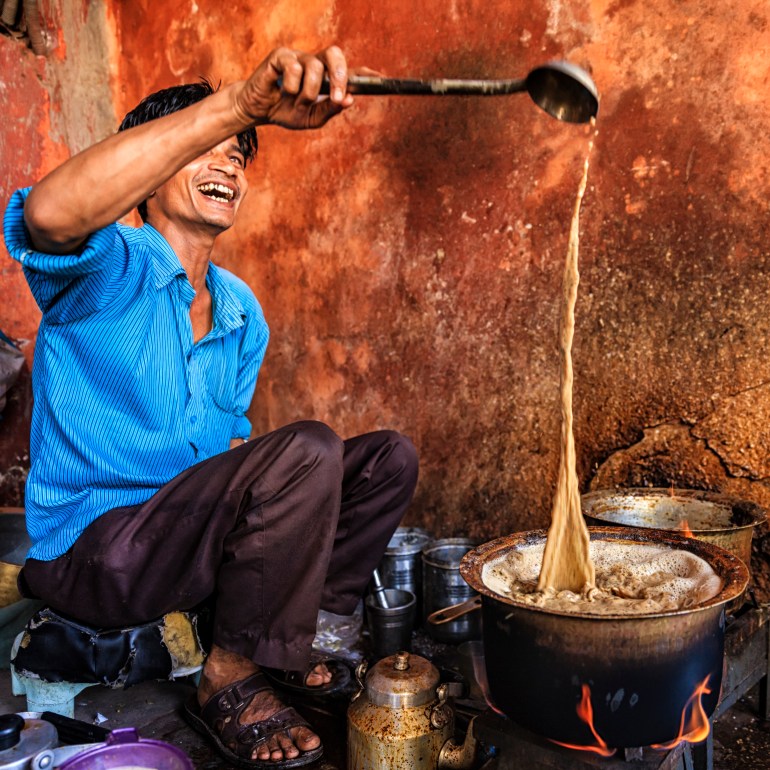 A laughing chai walla pours tea from a ladle held high back into the bubbling pot of chai