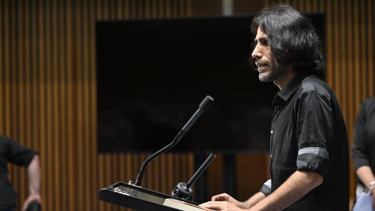 Behrouz Boochani speaking to Australian members of parliament. He is standing at a lectern and dressed in black.
