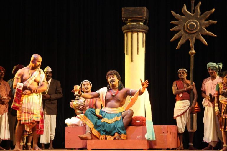 A photo of a group of people standing around on stage, in a play with a man sitting cross-legged in the middle.