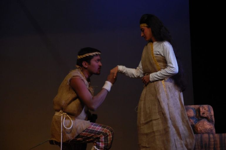 A photo of two people in a play. There's a man kneeling on the left holding the hand of a woman standing on the right.