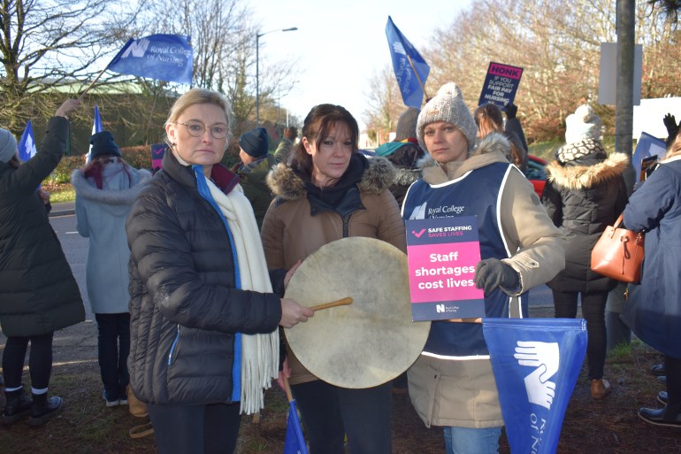 A photo of three people standing next to each other at a strike. The one on the left is holding a drum, covering the middle person's stomach and the one on the right is holding a sign with the words "staff shortages cost lives" on it.
