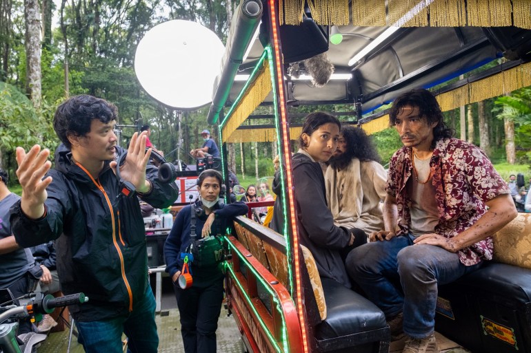 A scene from the Big Four. The director is explaining the scene on the left. A young woman and two men are sitting in the back of a carriage. It has cushioned seats and a fringed canopy. The clothes of the man in front are covered in blood. He is staring to one side of the frame.