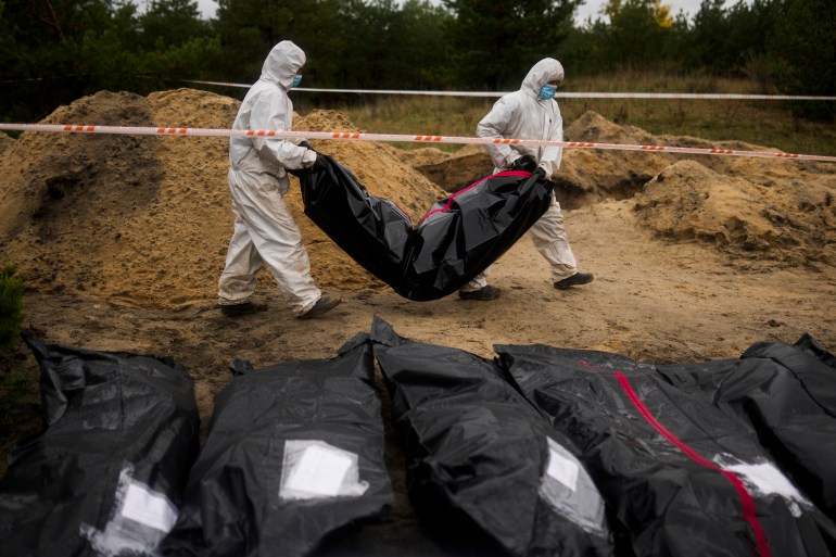 Two people in white-full-body suits carry a black, heavy-looking plastic bag while other black bags lie at the front of the photo