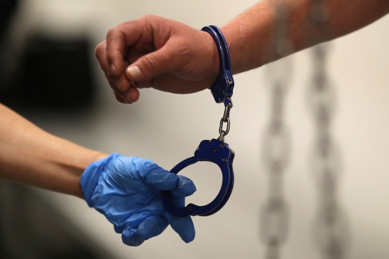 A blue-gloved hand holds one end of a pair of handcuffs. The other is around someone's wrist