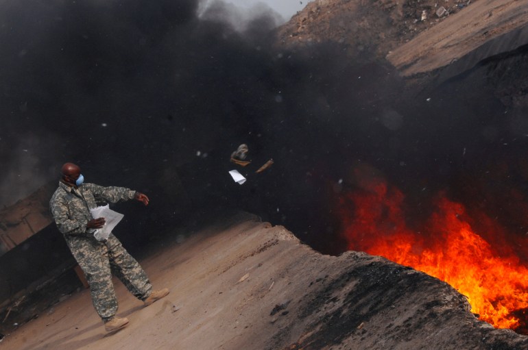 Black smoke rises from a fiery pit as a soldier tosses uniform material inside